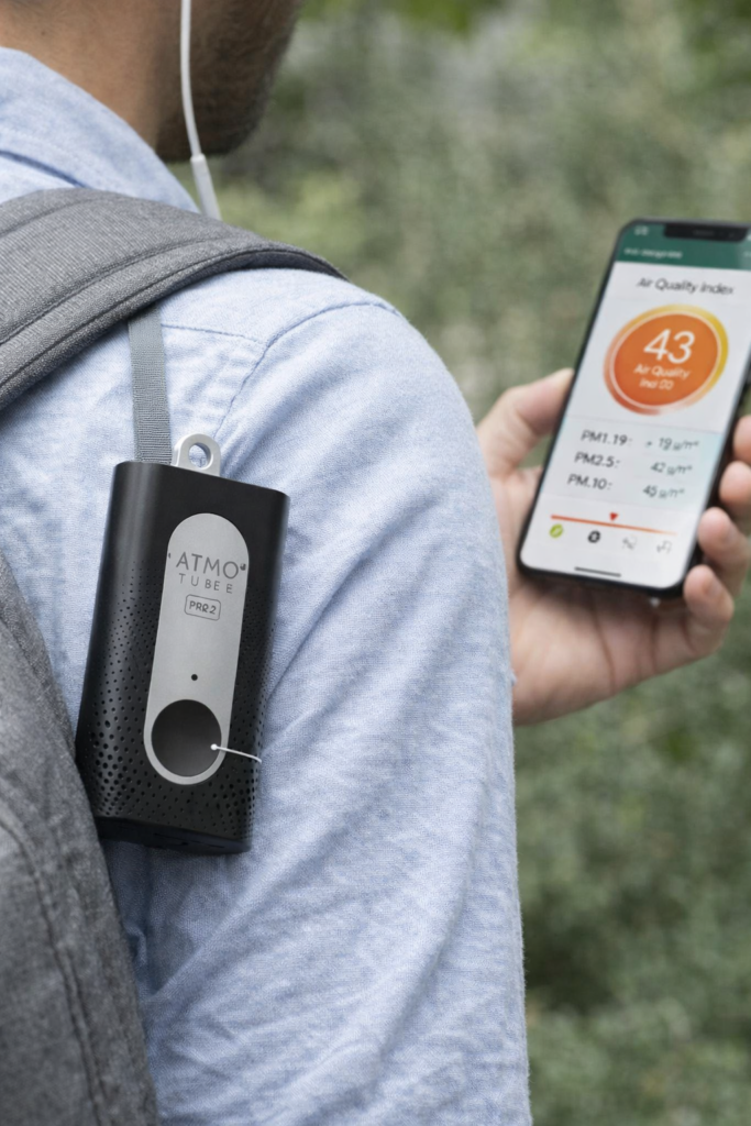 Person wearing the Atmotube PRO air quality monitor clipped to a backpack strap while viewing real-time particulate and air quality readings on a smartphone outdoors