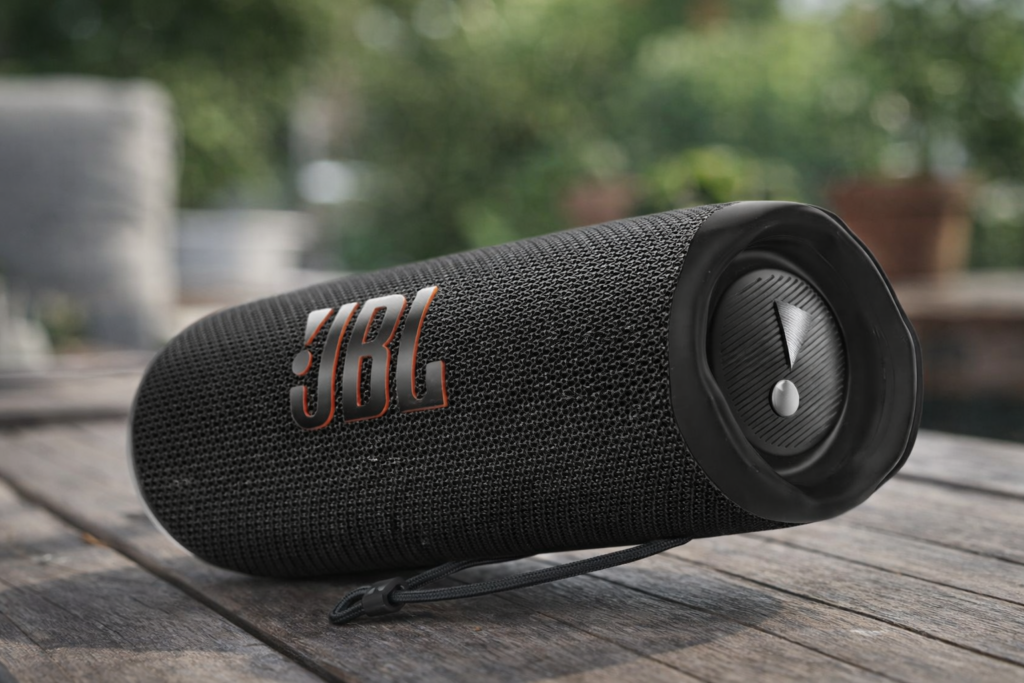 JBL Flip 6 portable Bluetooth speaker with cylindrical mesh design placed horizontally on a wooden table outdoors with blurred background